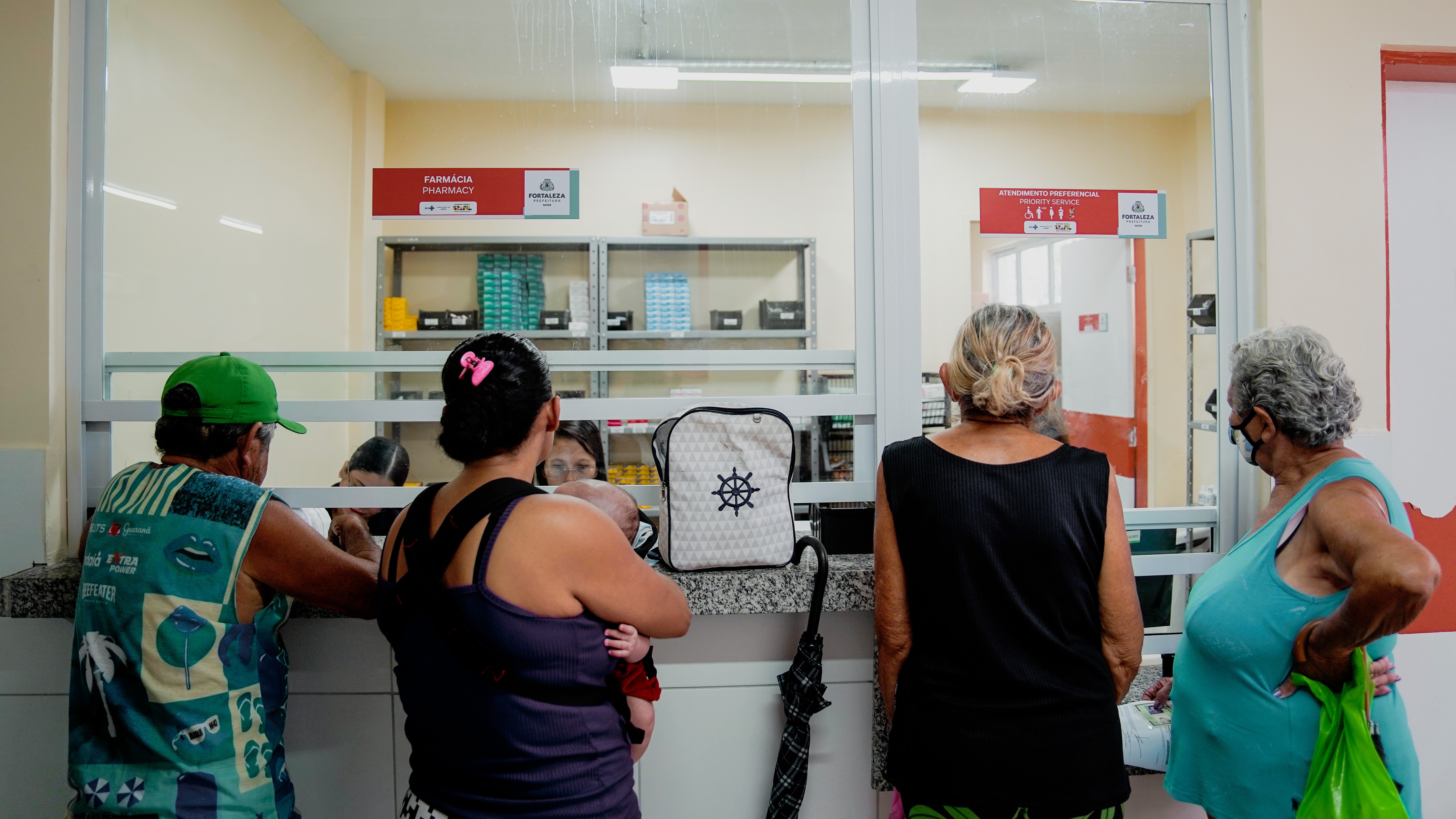 quatro pessoas de costas na frente do guichê de atendimento da farmácia de um posto de saúde